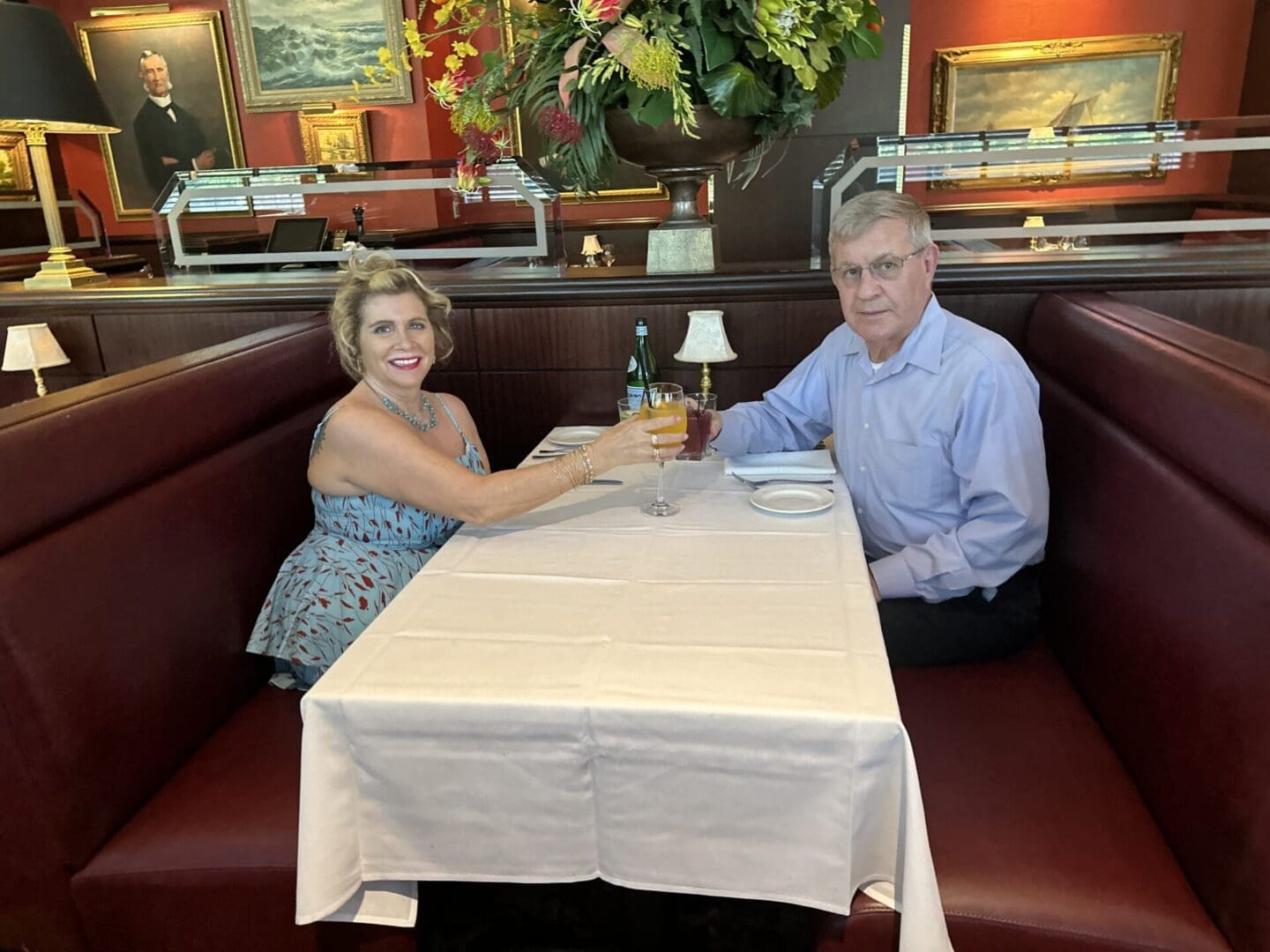 A man and woman sit at a booth in a restaurant, clinking glasses over a white tablecloth. A large floral arrangement and framed paintings are in the background.