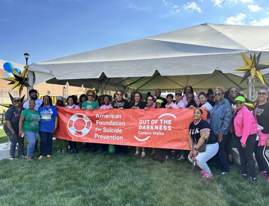 A group of people stand together holding a banner that reads "American Foundation for Suicide Prevention, Out of the Darkness Campus Walks" in front of a tent.