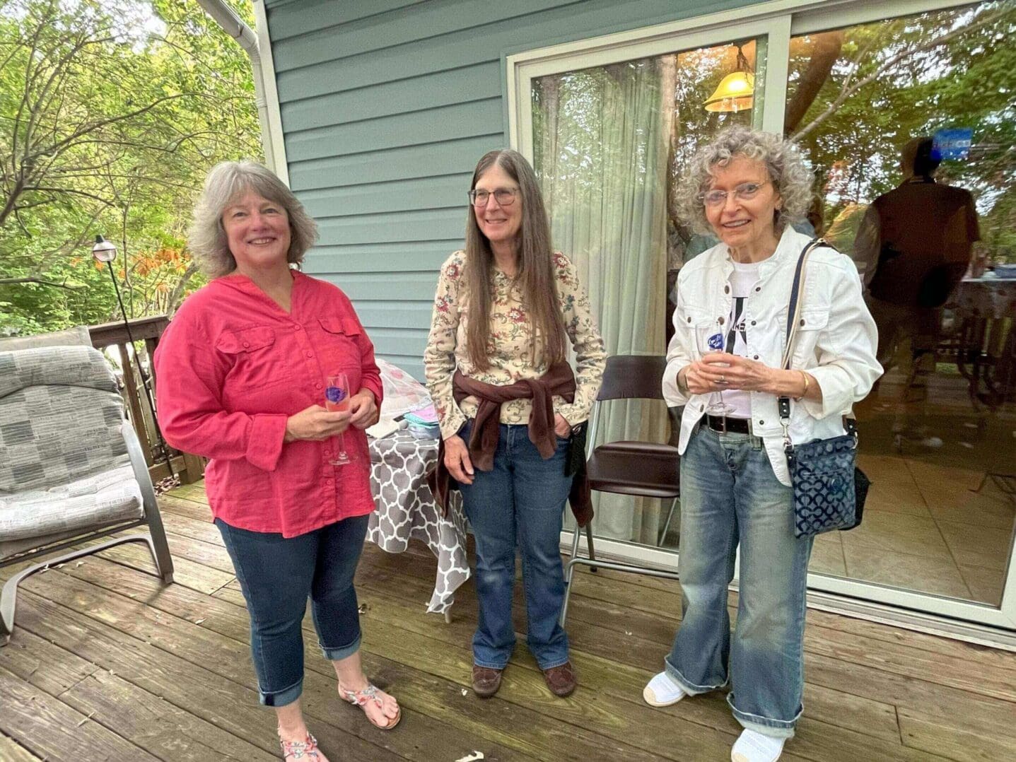 Three women smiling on a wooden porch outside a house.