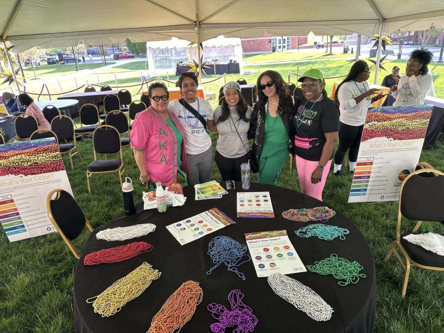 Four women standing behind a table with organized beads under a tent at an outdoor event.