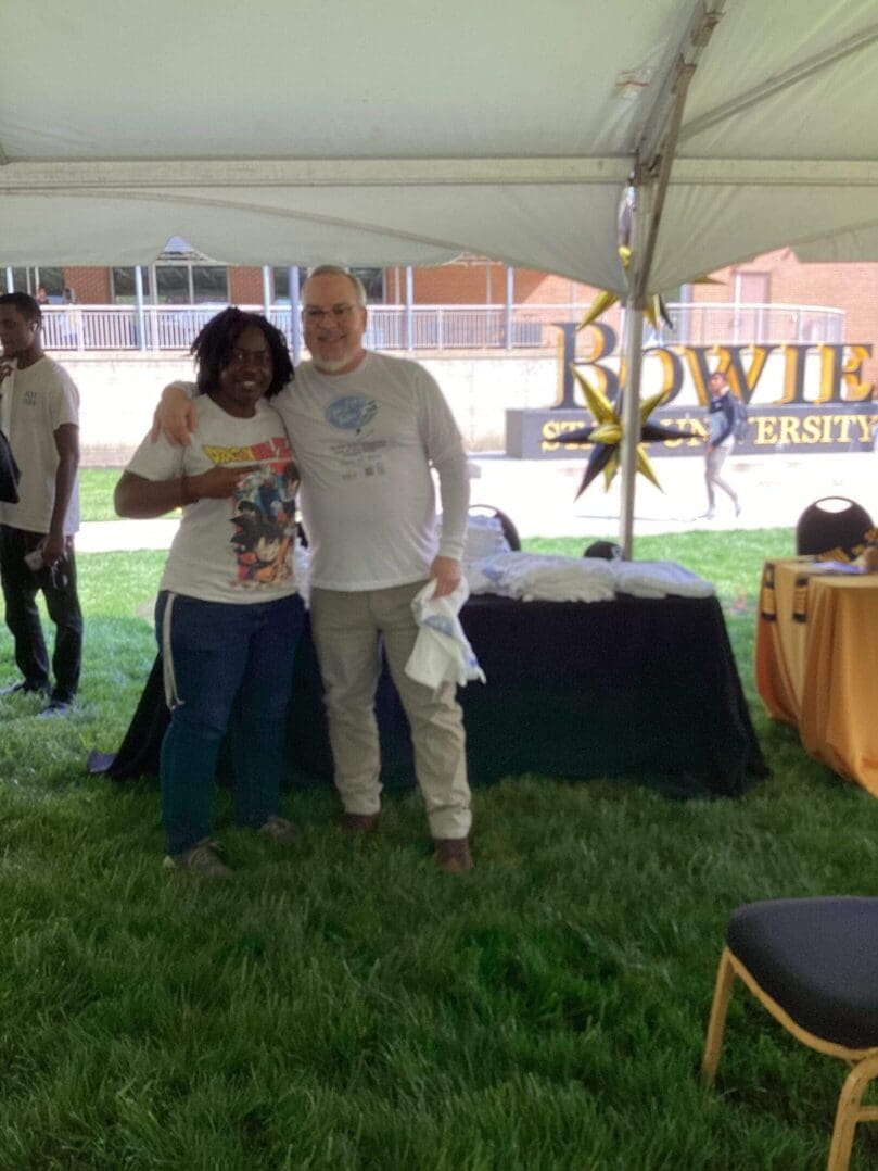 Two people posing under a tent at an outdoor event with a Bowie Festival sign.