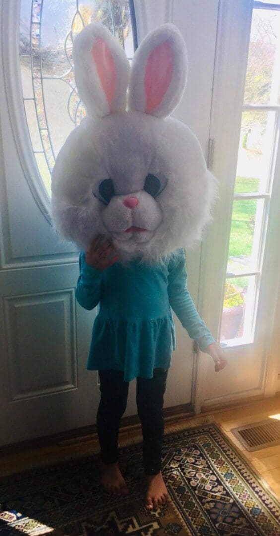 Child wearing oversized bunny head indoors.